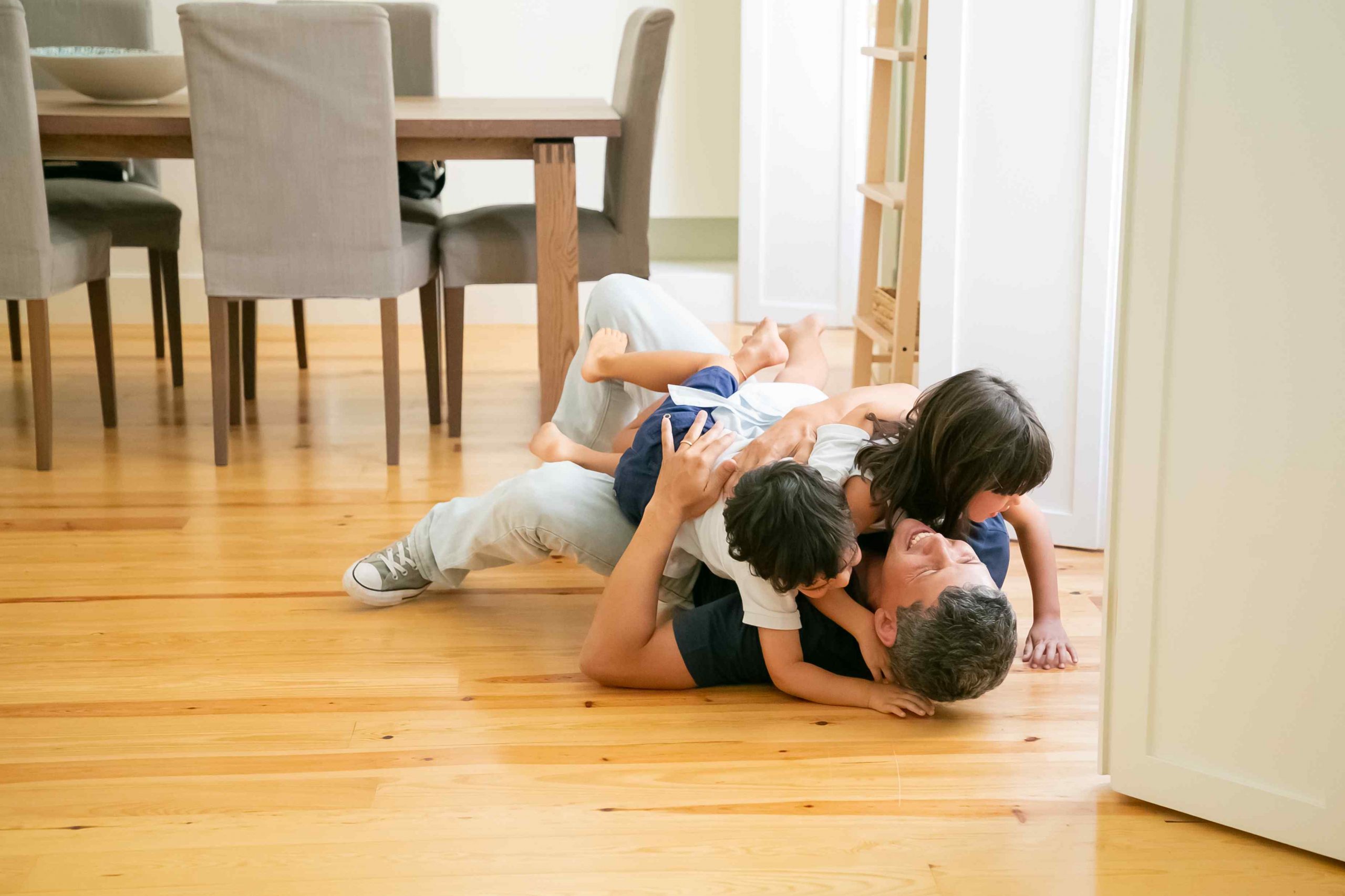 Laughing father lying on floor and hugging cute children. Happy attractive Caucasian dad embracing adorable kids, smiling and playing with son and daughter. Fatherhood, leisure and parenthood concept